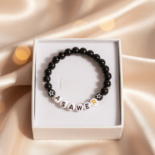 Black beaded bracelet with letter beads in a white box on a beige fabric background